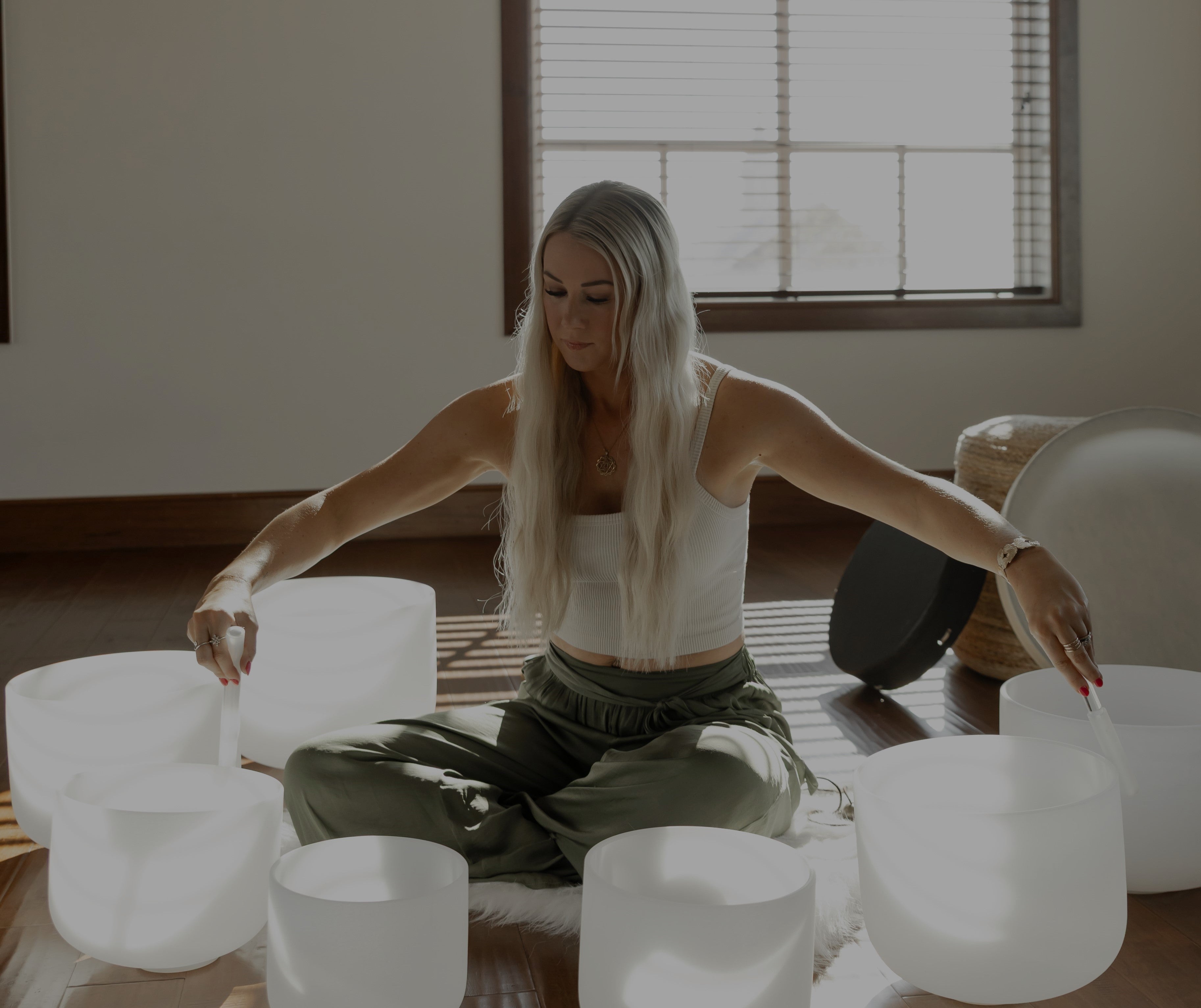 Woman sitting on the floor with crystal singing bowls in a room with a window.